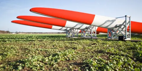 Drei Rotorblätter für Windkraftanlagen liegen auf einem Feld. Sie sind rot-weiß gestreift und warten auf ihren Einsatz nach dem EEG.