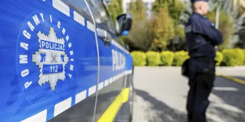 Policjant w mundurze stoi obok radiowozu z widocznym znakiem Policji i numerem 112.