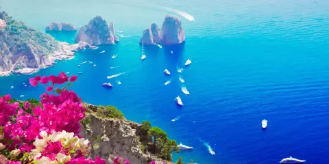 Un giorno a Capri: scogliere rocciose, mare blu intenso e barche che solcano le onde, con bouganville rosa in primo piano.