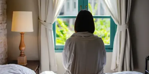 Osoba w szlafroku siedzi na łóżku, patrząc przez okno. Czy to objaw katatonii?