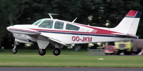 Eine weiße Beechcraft Bonanza mit roten und blauen Streifen hebt vom Gras ab.