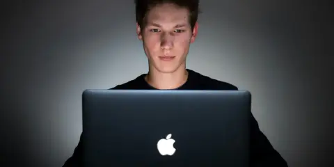 Młody mężczyzna pochylony nad laptopem, jego twarz oświetlona ekranem. Czy to początek uzależnienia od internetu?