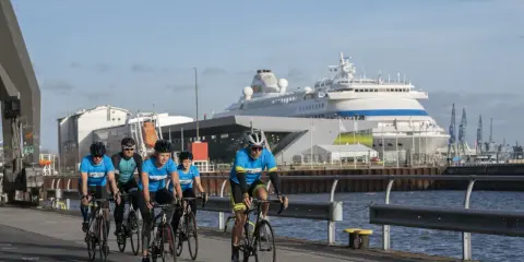 Fünf Radfahrer in blauen Trikots absolvieren ein Mini-Triathlon-Training am Hafen, ein großes Kreuzfahrtschiff im Hintergrund.