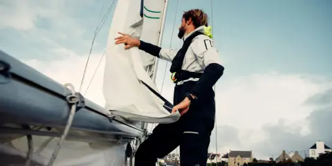 Un homme ajuste un type de voile sur un voilier, avec des maisons en arrière-plan.