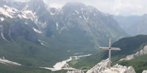 Kors på en klippig topp med en dal och snöklädda berg i bakgrunden, en vy över norra Albanien.