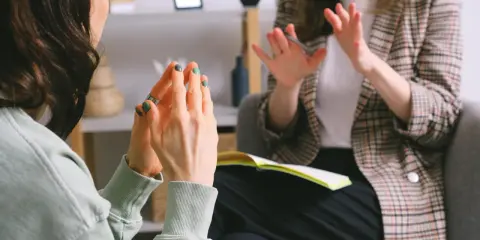 Kobieta z zielonymi paznokciami gestykuluje, jakby stresowała się byle czym. Obok niej siedzi druga osoba w kratkę.