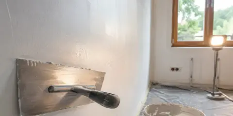 Narzędzie do wygładzania ścian, pokazujące jak położyć gładź. W tle wiadro z masą i okno.