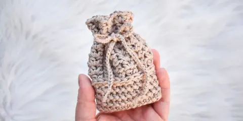 Monedero de ganchillo beige con cordón, sostenido en una mano sobre fondo blanco peludo.