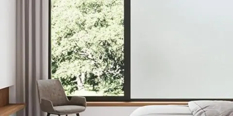 Habitación con cama, sillón y ventana con vidrio mate que filtra la luz del sol y la vegetación exterior.