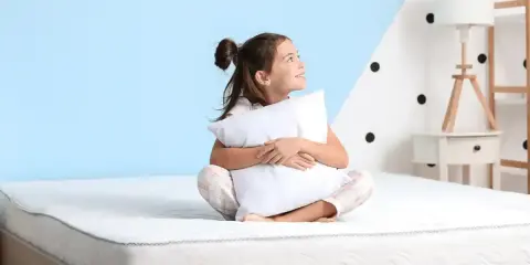 Niña sonriente abraza almohada en un colchón, pensando qué colchon es mejor para niños.