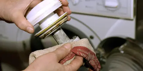 Hände reinigen das Flusensieb der Waschmaschine. Ein Tuch wischt Schmutz von dem ausgebauten Teil.