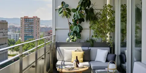 Acogedora terraza de una casa con sofá gris, plantas exuberantes y vistas urbanas. Ideal para relajarse al sol.