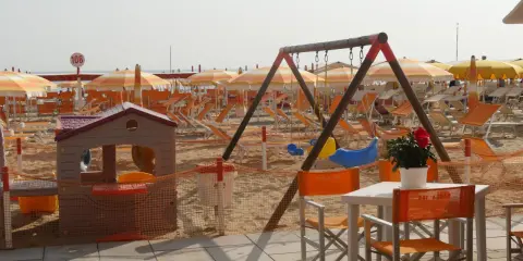 Area giochi e lettini sulla spiaggia 106 di Riccione, con ombrelloni arancioni e bianchi, altalena e casetta per bambini.