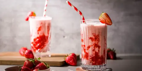 Zwei Gläser mit einem cremigen Erdbeer Milchshake, garniert mit frischen Erdbeeren und gestreiften Strohhalmen.