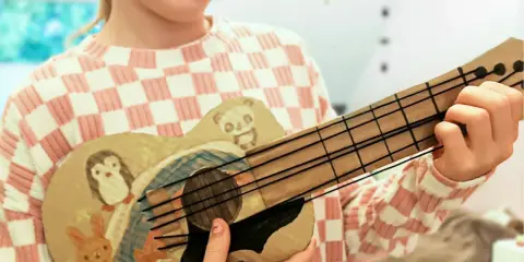 Kind bastelt Gitarre nach Vorlage. Die selbstgemachte Gitarre ist mit süßen Tieren wie Pinguin, Panda, Hase und Fuchs bemalt.