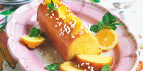 Torta al limone leggerissima, lucida e decorata con fettine d'arancia e zuccherini, servita su un piatto rosa con spicchi di limone e menta.