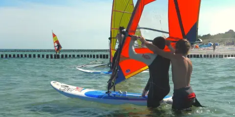 Zwei junge Männer bereiten sich auf das Windsurfen an der Ostsee vor. Ein weiterer Surfer ist in der Ferne zu sehen.