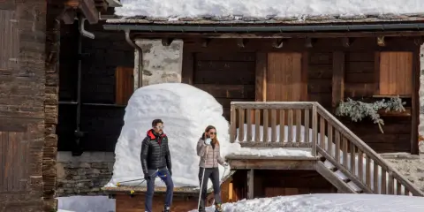 Coppia con racchette da neve a Riale Val Formazza, con un drone in volo sopra un tetto innevato.