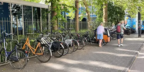 Viele Fahrräder stehen am Straßenrand, ein Mann mit Kind auf dem Arm und eine Frau mit Lastenrad gehen vorbei. Eine echte Stadt der kurzen Wege.