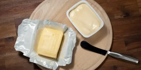 Zwei Butterpackungen, eine in Folie, die andere in einer weißen Schale, liegen auf einem Holzbrett. Eine leichte Butter weniger Fett für den gesunden Genuss.