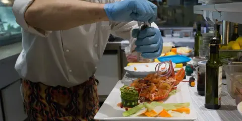 Chef con guanti blu macina pepe su un'aragosta, un piatto che sembra uscito da "lo zodiaco Rimini".