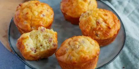 Fünf herzhafte Muffins mit Speck und Käse auf einem grauen Teller. Einer ist angeschnitten und zeigt die saftige Krume.
