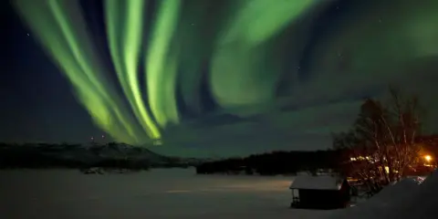 Grön aurora sweden dansar över ett snötäckt landskap med en liten stuga i förgrunden.