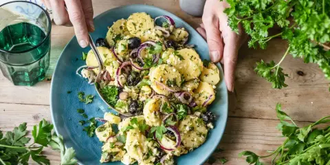 Ein köstlicher mediterraner Kartoffelsalat mit Oliven, roten Zwiebeln und Feta auf einem blauen Teller, garniert mit frischer Petersilie.