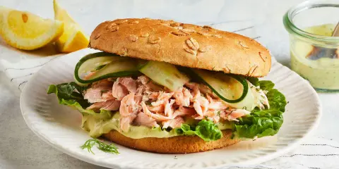 Ein Brötchen mit pulled Lachs, Salat, Gurkenscheiben und einer cremigen Sauce. Dazu Zitronenspalten und ein Dip.