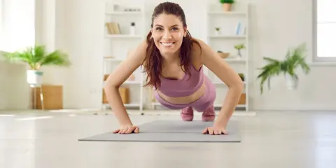 Frau in lila Sportkleidung macht Liegestütze für Yoga Muskelaufbau. Sie lächelt glücklich in die Kamera.