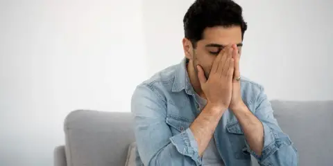 Mann sitzt auf Sofa, Hände vors Gesicht gepresst, Augen geschlossen. Fühlt sich nach dem Rauchen schwindelig.