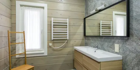 Salle de bain moderne avec murs en bois clair et pierre grise. Un meuble vasque en bois, un miroir, un radiateur et une échelle en bambou pour habiller un mur.