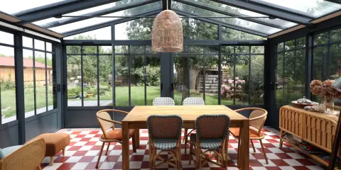 Salle à manger lumineuse dans une véranda avec toiture zinc. Table en bois, chaises colorées, sol à damier rouge et blanc, vue sur jardin verdoyant.