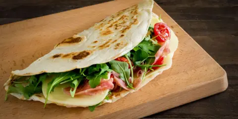 Piadina romagnola, un classico dal sapore autentico, ripiena di prosciutto, formaggio, rucola e pomodorini.