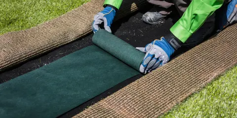 Manos enguantadas desenrollan césped artificial verde sobre una base negra. Paso clave para saber como colocar cesped artificial.