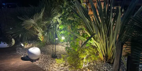 Jardin illuminé la nuit, créant une ambiance chaleureuse. La **norme éclairage extérieur** est respectée pour mettre en valeur la végétation luxuriante et le chemin en bois.