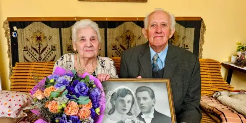 Para świętuje 70 rocznicę ślubu, trzymając zdjęcie z dnia zaślubin. W bukiecie kwiatów dominują fiolety i pomarańcze.