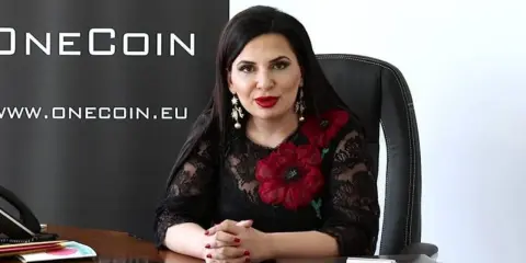 Rupert Ignatov, w czarnej koronkowej sukni z czerwonymi kwiatami, siedzi przy biurku.