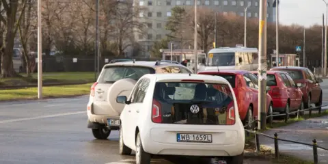 Biały Volkswagen up! zaparkowany tuż przy przejściu dla pieszych. W tle inne samochody i budynek.