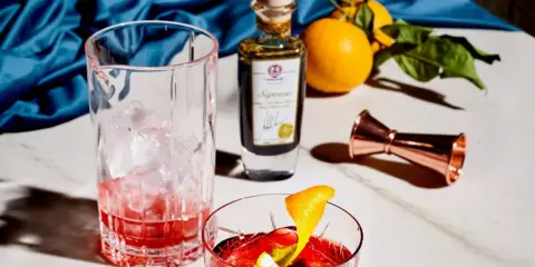 Ein Glas mit Eis und rotem Drink, daneben eine Flasche mit der Aufschrift "Saponoso". Orangen und ein Messbecher vervollständigen die Szene. Grappa, was ist das?