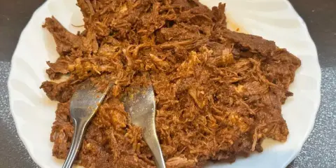 Delicioso pulled pork deshebrado en un plato blanco, listo para ser servido. Dos tenedores se clavan en la carne jugosa.