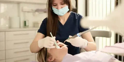 Dentystka w masce i rękawiczkach wykonuje zabieg u pacjentki w gabinecie stomatologicznym.