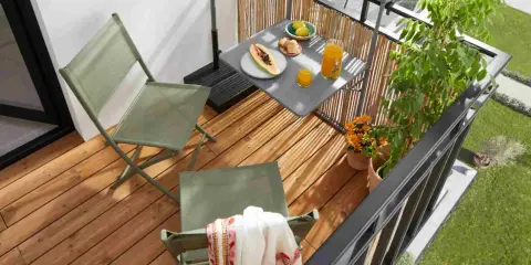 Idéal pour aménager un petit balcon en longueur : deux chaises pliantes vertes, une table avec fruits et jus, et des plantes.