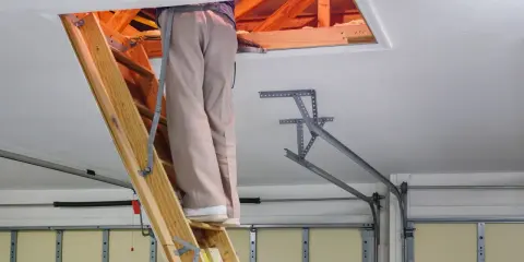 Osoba wspina się po drewnianej drabinie do włazu na strych. Widać drewniane belki stropowe i izolację. To dobry przykład, jak wykończyć właz na strych.