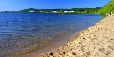 Słoneczny dzień nad zalewem Pilica. Piaszczysta plaża, spokojna woda i zielone wzgórza w tle. Kilka osób pływa i odpoczywa.
