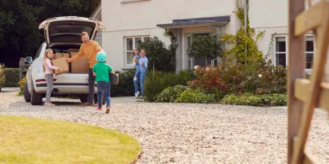 Rodzina rozładowuje samochód na podjeździe z kruszywa. Ojciec z dziećmi wyjmuje kartony, mama z córką wita ich przed domem.