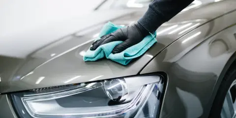 Ręka w czarnej rękawiczce z turkusową mikrofibrą wykonuje polerowanie lakieru samochodowego, nadając mu lustrzany połysk.
