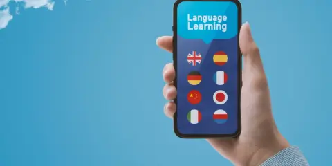 Dłoń trzyma smartfon z aplikacją do nauki języków. Wybór między Busuu czy Duolingo? Na ekranie flagi: UK, Hiszpanii, Niemiec, Francji, Chin, Japonii, Włoch, Polski.