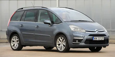 Szary Citroen Picasso z napisem "Picasso" na błotniku, zaparkowany przed budynkiem.