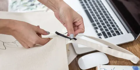 Ręce tworzą torbę DIY, przyszywając czarną wstążkę do płóciennego materiału. Obok leży laptop i myszka.